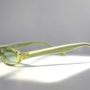 K&ouml;nnte beinhalten: Hellgelbe Cat-Eye-Sonnenbrille mit gr&uuml;n verlaufenden Gl&auml;sern. Die Fassungen und B&uuml;gel sind von transluzenter, hellgelber Farbe. Die Sonnenbrille ist leicht angewinkelt und wirft einen Schatten auf die wei&szlig;e Oberfl&auml;che.