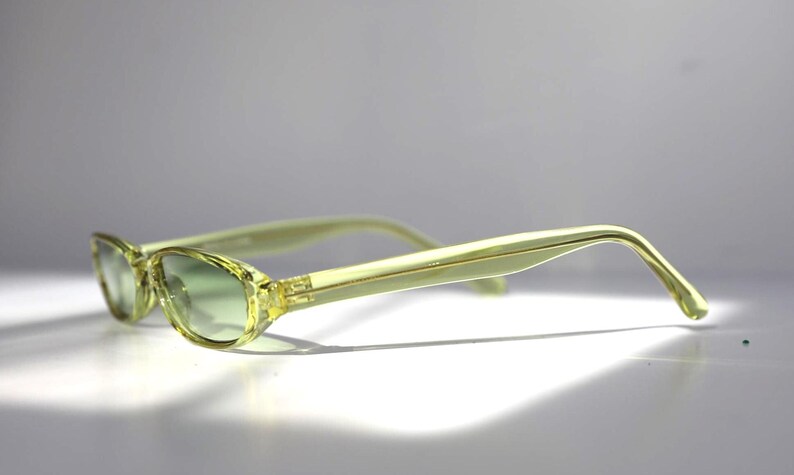 K&ouml;nnte beinhalten: Hellgelbe Cat-Eye-Sonnenbrille mit gr&uuml;n verlaufenden Gl&auml;sern. Die Fassungen und B&uuml;gel sind von transluzenter, hellgelber Farbe. Die Sonnenbrille ist leicht angewinkelt und wirft einen Schatten auf die wei&szlig;e Oberfl&auml;che.