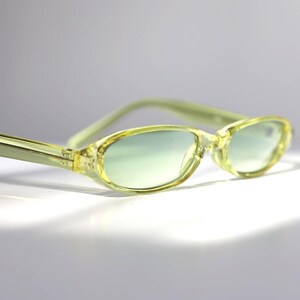 K&ouml;nnte beinhalten: Hellgelbe ovale Sonnenbrille mit leicht gr&uuml;nlichem Farbton. Die Fassungen und B&uuml;gel sind aus durchscheinendem Gelb. Die Gl&auml;ser haben einen Farbverlauf, der von Gr&uuml;n oben nach transparent unten verl&auml;uft. Die Sonnenbrille ist vor einem wei&szlig;en Hintergrund platziert.