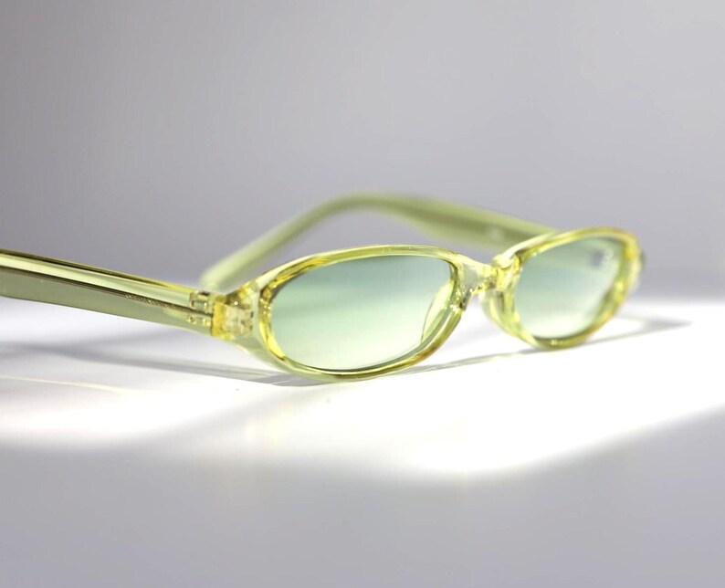 K&ouml;nnte beinhalten: Hellgelbe ovale Sonnenbrille mit leicht gr&uuml;nlichem Farbton. Die Fassungen und B&uuml;gel sind aus durchscheinendem Gelb. Die Gl&auml;ser haben einen Farbverlauf, der von Gr&uuml;n oben nach transparent unten verl&auml;uft. Die Sonnenbrille ist vor einem wei&szlig;en Hintergrund platziert.