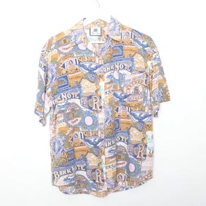 May include: Short-sleeved button-down shirt with a repeating pattern of vintage banknote designs in shades of blue, tan, and pink. The shirt has a classic collar and a single chest pocket. The shirt is hanging on a white hanger.