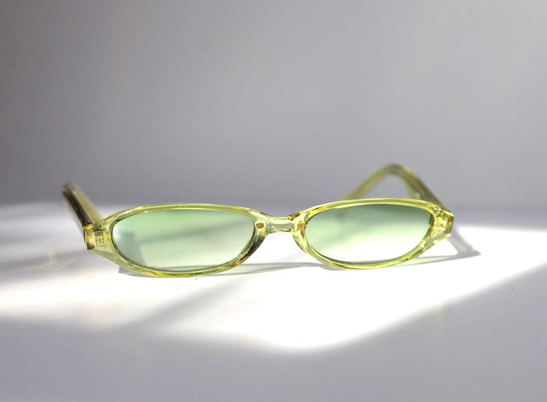 K&ouml;nnte beinhalten: Hellgr&uuml;ne ovale Sonnenbrille mit passenden Rahmen. Die Gl&auml;ser haben einen dezenten Gr&uuml;nton, und die Rahmen sind eine durchscheinende, hellgr&uuml;ne Farbe. Die Sonnenbrille ist vor einem wei&szlig;en Hintergrund platziert.