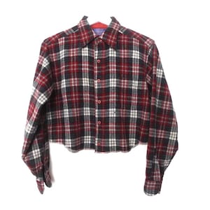 Peut inclure: Chemise en flanelle courte à carreaux rouges, noirs et blancs. La chemise à manches longues a une fermeture boutonnée sur le devant, un col et des poignets boutonnés. La chemise est suspendue sur un fond blanc.