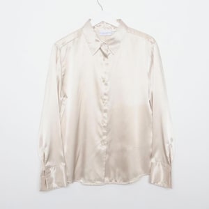 Peut inclure: Chemise à manches longues de couleur champagne clair, avec une fermeture boutonnée et un col et des poignets classiques. La chemise est suspendue à un cintre blanc sur fond blanc. Le tissu a un éclat subtil.