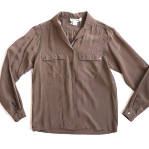 May include: A long-sleeved, button-down shirt in a muted brown color. The shirt features two chest pockets with button closures and a classic collar. The fabric has a subtle sheen, suggesting a silky texture. The shirt is laid flat on a white surface.