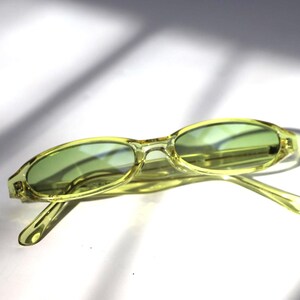 K&ouml;nnte beinhalten: Ovale, hellgr&uuml;ne Sonnenbrille mit passenden get&ouml;nten Gl&auml;sern. Die Fassungen sind eine durchscheinende, blassgelb-gr&uuml;ne Farbe. Die Sonnenbrille liegt vor einem wei&szlig;en Hintergrund mit Schatten.