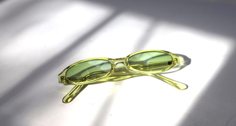 K&ouml;nnte beinhalten: Ovale, hellgr&uuml;ne Sonnenbrille mit passenden get&ouml;nten Gl&auml;sern. Die Fassungen sind eine durchscheinende, blassgelb-gr&uuml;ne Farbe. Die Sonnenbrille liegt vor einem wei&szlig;en Hintergrund mit Schatten.