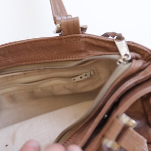 May include: A brown leather handbag with a top zipper closure, featuring a beige fabric interior lining, a zippered inner pocket, and two leather handles attached with metal hardware. The bag has a structured opening and visible stitching.