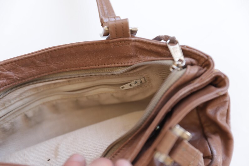May include: A brown leather handbag with a top zipper closure, featuring a beige fabric interior lining, a zippered inner pocket, and two leather handles attached with metal hardware. The bag has a structured opening and visible stitching.