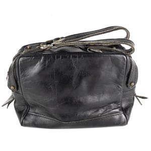 Puede incluir: Bolso de mano de cuero negro con asa superior y bandolera. El bolso tiene forma rectangular con un aspecto ligeramente desgastado. Los herrajes son plateados y el bolso parece usado.
