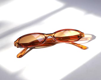Könnte beinhalten: Ovale Sonnenbrille mit einem durchscheinenden orangefarbenen Rahmen und getönten Gläsern. Die Sonnenbrille liegt auf einer weißen Oberfläche, mit Schatten, die auf den Hintergrund geworfen werden. Die Gesamtästhetik ist minimalistisch und stilvoll.
