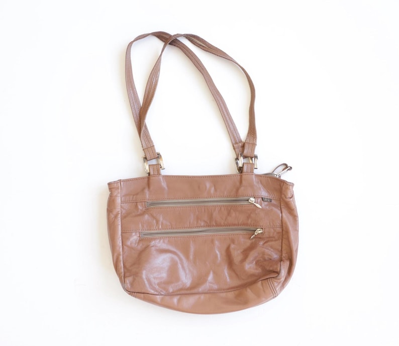 May include: A medium-sized brown leather shoulder bag featuring two horizontal zippered pockets on the front, a main top zipper closure, and two thin shoulder straps attached with silver-toned metal hardware connectors at the top.