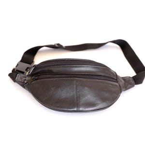 May include: A black leather fanny pack featuring two horizontal zippered compartments on the front. The bag includes an adjustable black nylon strap with a plastic side-release buckle, providing a secure closure for carrying small personal items.