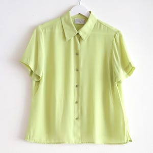 May include: A light green short-sleeved button-down shirt hanging on a white hanger. The shirt has a classic collar and buttons down the front. The fabric appears to be lightweight and slightly sheer, with a subtle sheen. The shirt is a solid, pale green color.