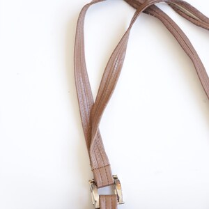 May include: A brown leather handbag featuring two thin, textured shoulder straps attached to the main body with rectangular silver-toned metal hardware. The bag has a smooth, consistent brown finish and visible stitching along the strap edges.