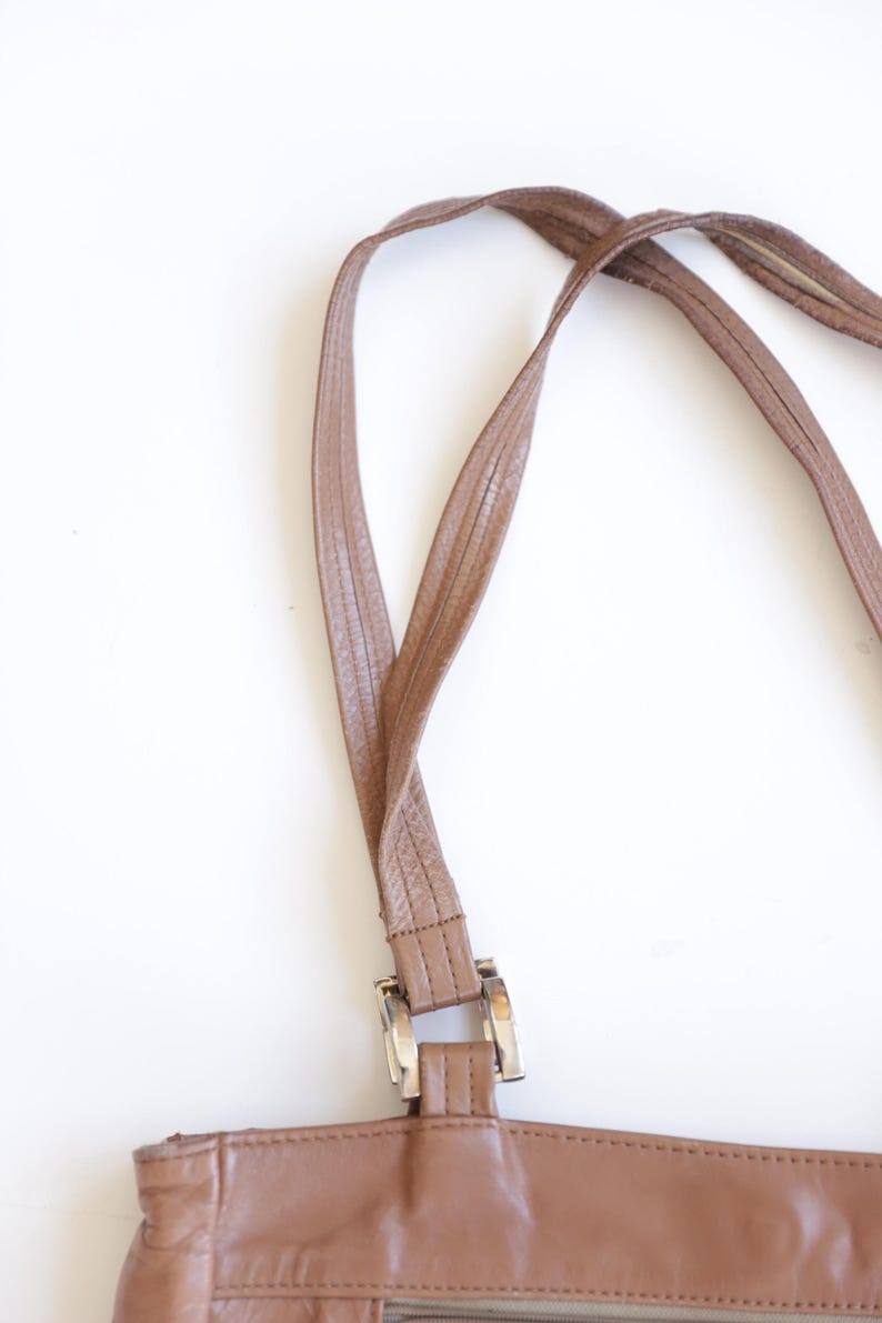 May include: A brown leather handbag featuring two thin, textured shoulder straps attached to the main body with rectangular silver-toned metal hardware. The bag has a smooth, consistent brown finish and visible stitching along the strap edges.