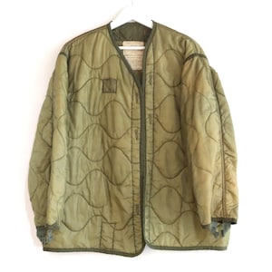 May include: Olive green quilted jacket with a round collar and button closure. The jacket has a wavy quilted pattern and is hanging on a white hanger. The jacket has a label on the inside collar.