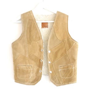 May include: A tan suede leather vest featuring a patchwork design with visible stitching. The garment has a white faux shearling lining, a V-neckline, three silver-toned metal snap buttons on the front, and two patch pockets at the waist.