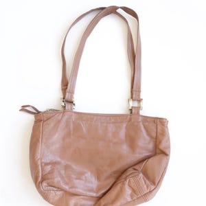 May include: A medium-sized, tan leather shoulder bag with a soft, slouchy silhouette. It features two thin shoulder straps attached with silver-toned rectangular metal hardware, a top zipper closure with two pull tabs, and visible stitching.