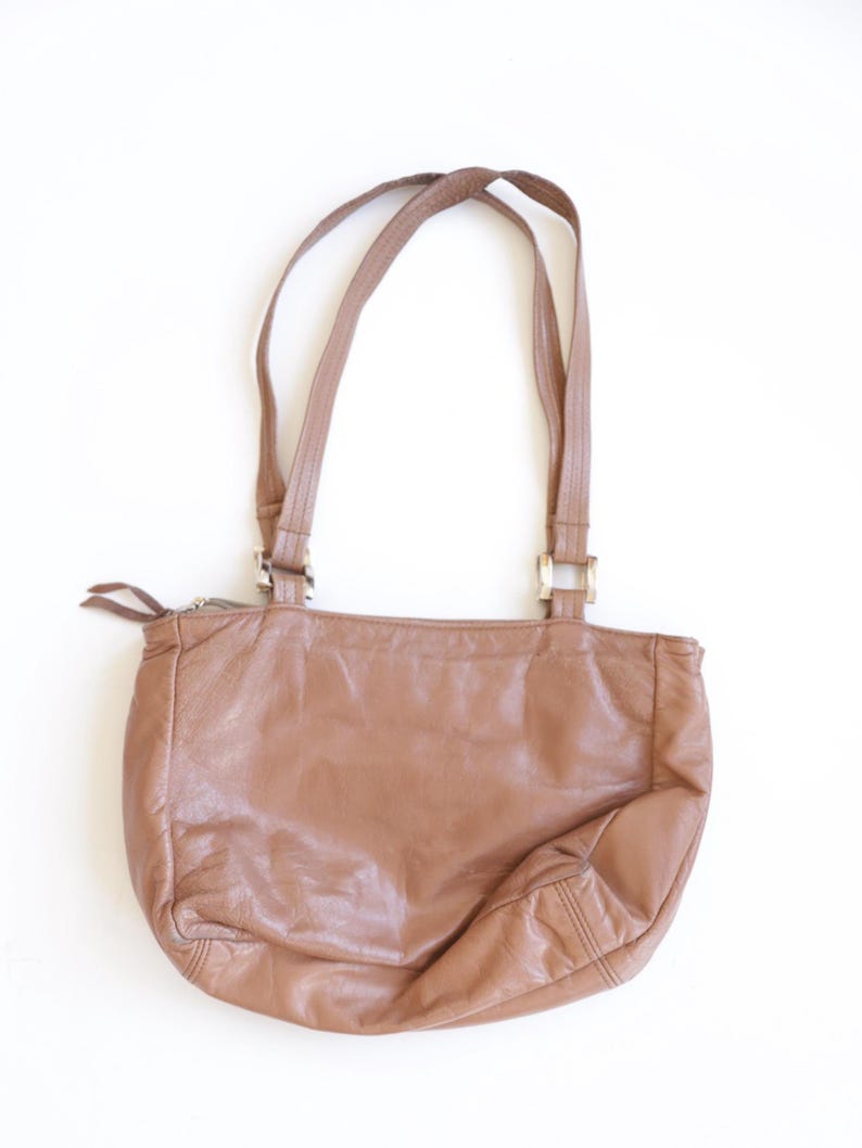 May include: A medium-sized, tan leather shoulder bag with a soft, slouchy silhouette. It features two thin shoulder straps attached with silver-toned rectangular metal hardware, a top zipper closure with two pull tabs, and visible stitching.