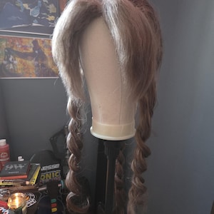 May include: A light brown wig with long, braided pigtails is displayed on a mannequin head. The wig has a natural-looking hairline and is styled with voluminous waves and curls. The mannequin is mounted on a black stand.