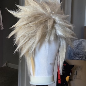 May include: A light blonde, spiky wig styled on a white mannequin head. The wig has a textured, layered appearance with a few strands hanging down. A silver pin secures the wig. The mannequin is on a black stand.