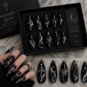 May include: A set of black press-on nails in a box labeled Midnight Ritual. The nails feature silver metallic accents including crosses, crescent moons, butterflies, roses, and stars. The box includes text reading Not just nails, but a piece of your story.