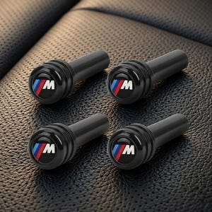 May include: Four black valve stem caps with a circular emblem featuring the letter "M" in white, with red and blue stripes. The caps are designed for automotive use and are displayed on a textured surface.