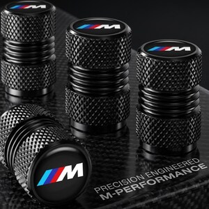 Könnte beinhalten: Schwarze Reifenventilkappen mit strukturiertem Design. Jede Kappe zeigt das BMW M-Logo in Blau, Rot und Weiß auf schwarzem Hintergrund. Die Kappen sind auf einer dunklen Oberfläche mit dem Text "PRECISION ENGINEERED M-PERFORMANCE" angeordnet.