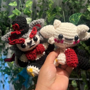 May include: Two handmade crochet dolls. One doll is gray, black, and red, wearing a top hat and bow tie. The other doll is white with red accents. Both dolls have black button eyes and are held in a hand.