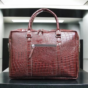 Puede incluir: Un maletín de cuero burdeos con textura de piel de cocodrilo. El bolso tiene un asa superior, un bolsillo frontal con cremallera y herrajes plateados. El bolso es rectangular y parece resistente.