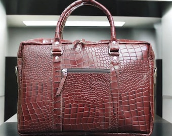 May include: A burgundy leather briefcase with a crocodile skin texture. The bag has a top handle, a zippered front pocket, and silver-toned hardware. The bag is rectangular and appears to be sturdy.