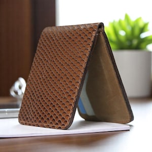 Op de afbeelding: Een bruine leren portemonnee met een textuurpatroon dat op schubben lijkt. De portemonnee is open en onthult een glimp van de binnenkant en een paar kaarten. De portemonnee ligt op een wit oppervlak.