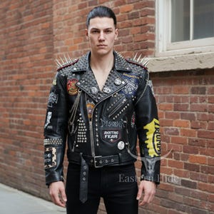 Puede incluir: Una chaqueta de moto de cuero negro con pinchos metálicos plateados en los hombros, numerosos remaches circulares y varios parches bordados con textos como Disorder, Awaiting Fear, Michelin y Triumph, que incluye un cinturón en la cintura.