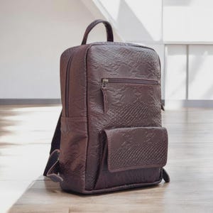 May include: A burgundy backpack with a textured, snakeskin-like pattern. The bag has a top handle, a front pocket, and a zippered compartment. The backpack is standing on a wooden floor.