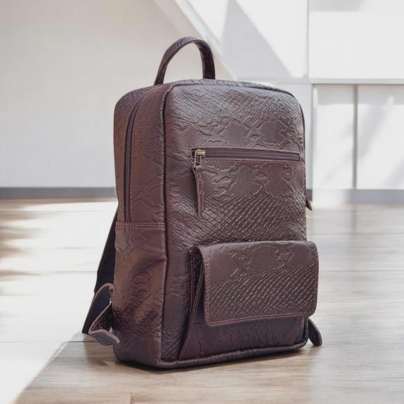 May include: A burgundy backpack with a textured, snakeskin-like pattern. The bag has a top handle, a front pocket, and a zippered compartment. The backpack is standing on a wooden floor.