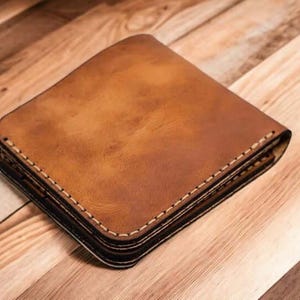 Puede incluir: Una cartera de cuero marrón con costuras blancas. La cartera es cuadrada y parece estar doblada. El cuero tiene un aspecto envejecido, con variaciones de color. La cartera está sobre una superficie de madera.