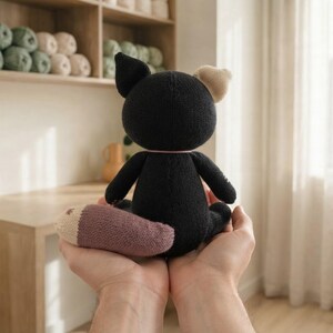 Op de afbeelding: Een gebreide knuffel in de vorm van een kat met zwarte vacht, een lichtbeige oor en een lange staart met een lichtbeige punt. Het speelgoed heeft een ronde kop, kleine oren en een zachte textuur, weergegeven vanaf de achterkant.