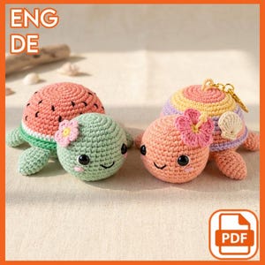 Puede incluir: Juguetes de tortuga de ganchillo en varios colores. Uno tiene temática de sandía, otro es verde con una flor rosa, y el tercero es melocotón con una flor rosa y un llavero. Las tortugas tienen caras sonrientes.
