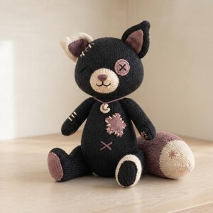 Op de afbeelding: Een gebreid pluche kattenspeeltje in zwart met een cr&egrave;mekleurige snuit en binnenkant van de oren. Het heeft een mauve stervormige patch op de borst, een mauve voetzooltje, een mauve maanhanger aan een koordje en een mauve staart met stersteek.
