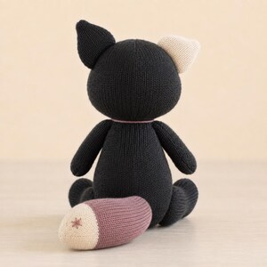 Op de afbeelding: Een gebreid knuffeldier in de vorm van een kat, voornamelijk zwart met een cr&egrave;mekleurig oor en een staart met een mauve gedeelte en een cr&egrave;mekleurige punt met een klein stervormig stiksel. Het speelgoed is van achteren in zittende houding.
