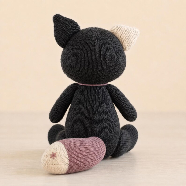 Op de afbeelding: Een gebreid knuffeldier in de vorm van een kat, voornamelijk zwart met een cr&egrave;mekleurig oor en een staart met een mauve gedeelte en een cr&egrave;mekleurige punt met een klein stervormig stiksel. Het speelgoed is van achteren in zittende houding.