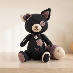 Op de afbeelding: Een zwart gebreid pluche kattenspeeltje met een beige snuit, &eacute;&eacute;n knoopoog en &eacute;&eacute;n geborduurd oog. Het heeft een mauve stervormige patch op de borst, een maansikkelhanger aan een dun koordje en een groot, losstaand mauve en beige staartstuk.