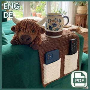 May include: A brown crocheted sofa armrest with two white pockets, holding a remote and a phone. A crocheted Highland cow and a floral mug with a white and brown cozy sit on the armrest. The armrest is on a green sofa.