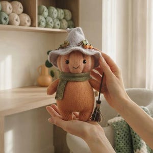Könnte beinhalten: Eine gestrickte Kürbispuppe mit einem hellvioletten Hexenhut, der mit kleinen orangefarbenen Kürbissen verziert ist, und einem grünen Schal. Die Puppe hat schwarze Perlenaugen, einen aufgestickten Mund und hält einen Miniaturbesen aus Zweigen.
