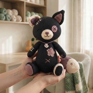 May include: A black knitted plush cat toy featuring a cream-coloured muzzle, one button eye, and one stitched eye. The toy has mauve-coloured accents on the ears, paw pads, and a star-shaped patch on the chest, with a small crescent moon charm necklace.