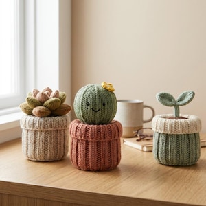 May include: Three knitted plant decorations in ribbed pots. The left is a multi-toned succulent, the center is a round green cactus with a small yellow flower and a stitched face, and the right is a green sprout with two leaves in a cream pot.