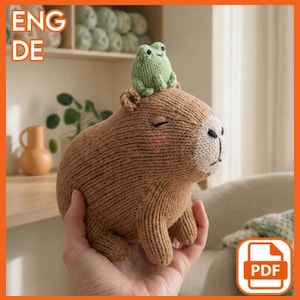 Könnte beinhalten: Ein handgefertigtes, gestricktes Capybara-Plüschtier in Brauntönen, mit einem kleinen grünen Frosch auf dem Kopf. Das Capybara hat eine weiße Schnauze, geschlossene Augen und rosa Wangen. Der Text "ENG DE" und ein PDF-Symbol sind enthalten.