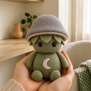 May include: A small, knitted plush toy shaped like a mushroom creature. It features a grey cap, a sage green body, and two black bead eyes. A white crescent moon shape is stitched onto the center of the chest. The toy has a rounded head and body.