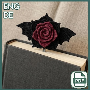 Op de afbeelding: Handgemaakte gehaakte boekenlegger met een donkerrode roos in het midden, omringd door zwarte vleermuisvleugelvormige bladeren. De boekenlegger is geplaatst in een hardcover boek met een donkergrijze kaft. De tekst "ENG DE" is zichtbaar.